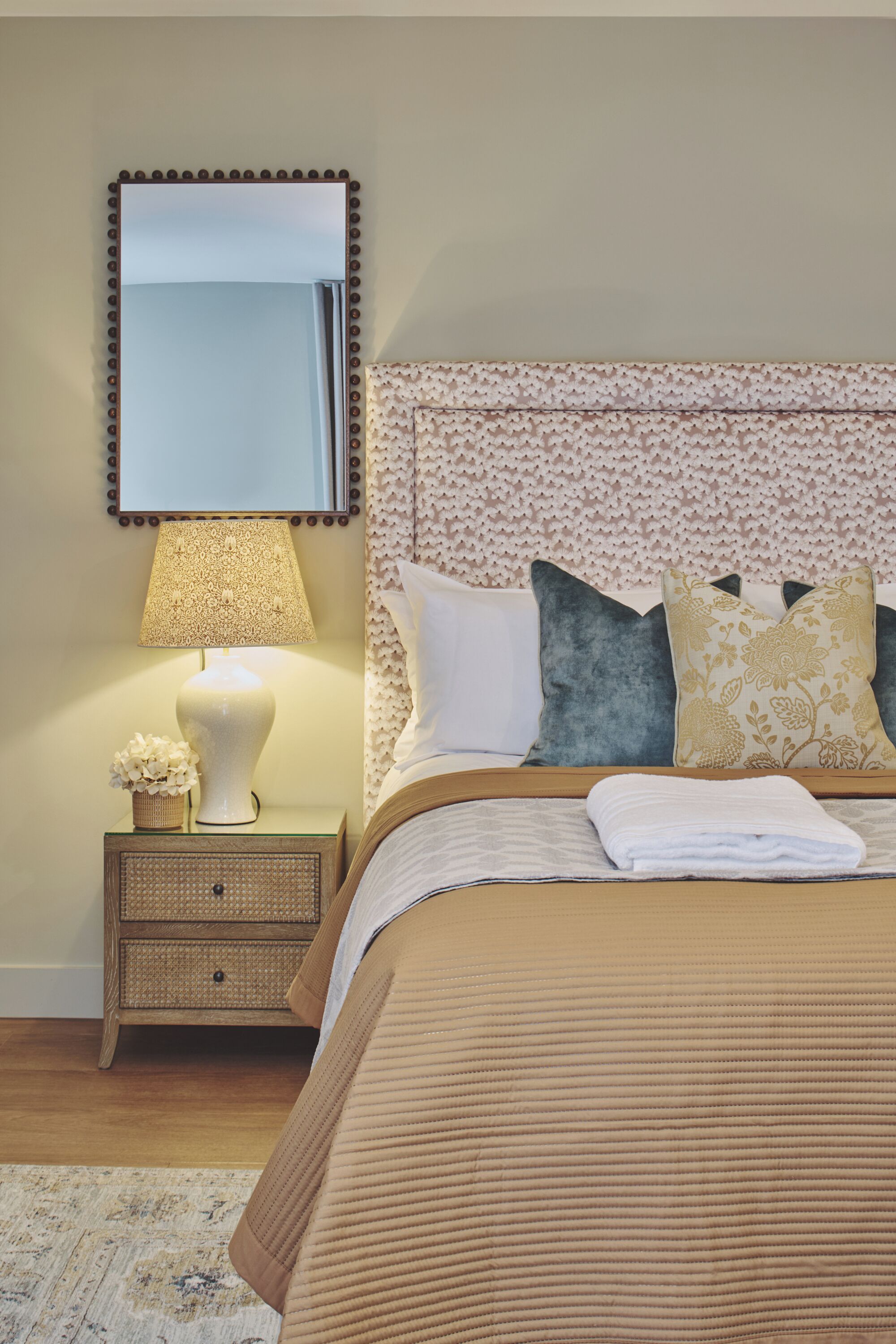 A neatly made bed with a patterned headboard, a bedside lamp, and a small wicker nightstand with a decorative item.