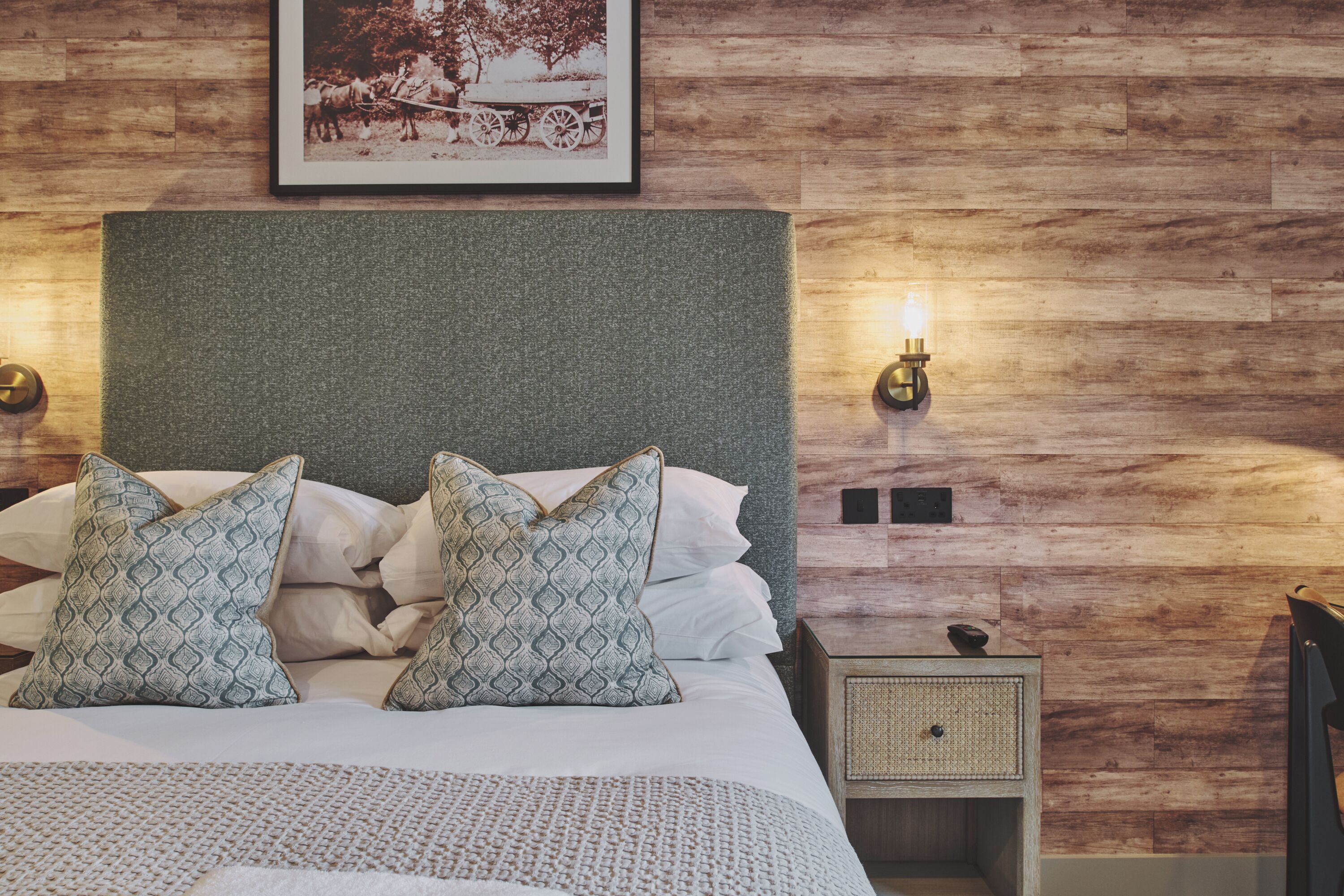 A neatly made bed with patterned pillows, a headboard, a bedside lamp, and a framed picture on the wall.