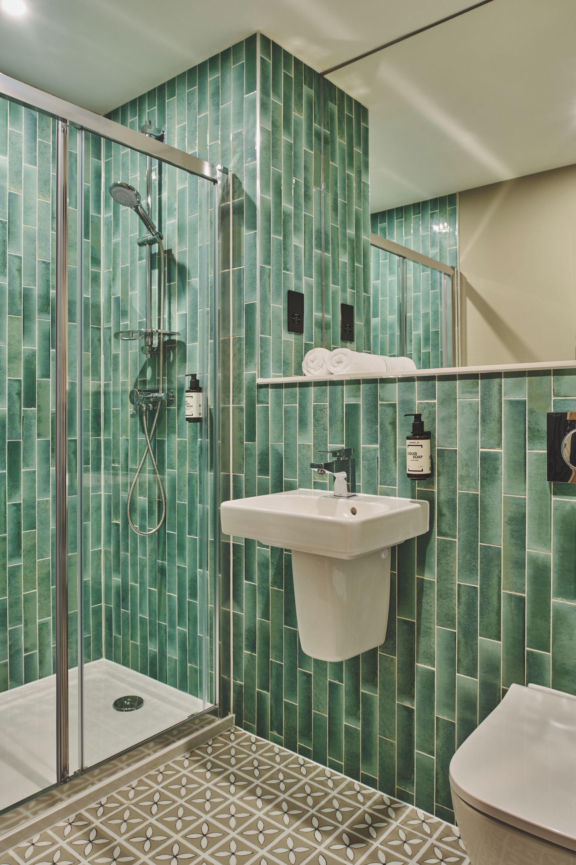 A bathroom with green tiled walls, a glass shower enclosure, a white sink, and a toilet.