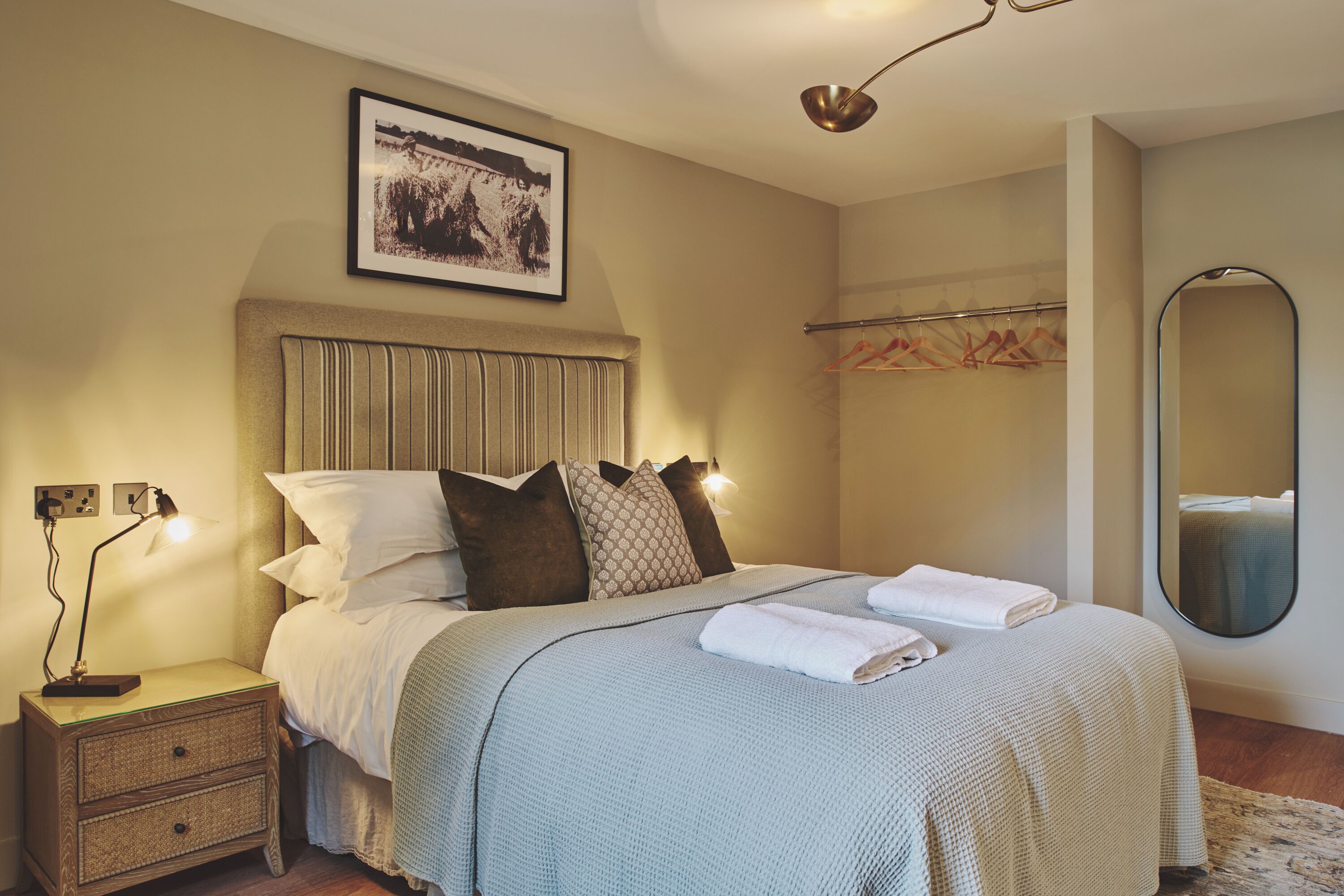 A neatly made bed with a headboard, a bedside table with a lamp, a framed picture on the wall, and a mirror on the wall.