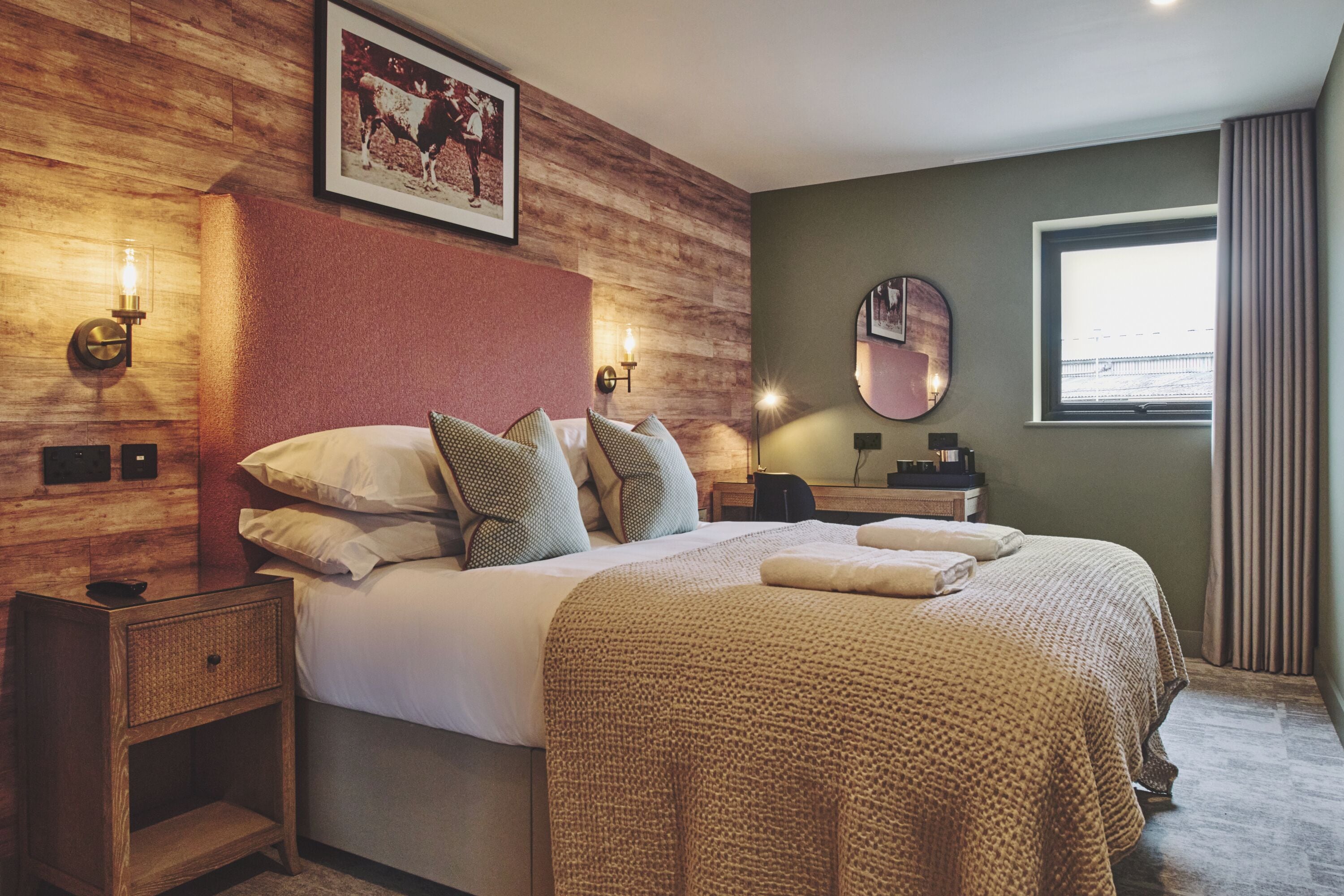 Une chambre d’hôtel avec une tête de lit en bois, un grand lit avec de nombreux oreillers, des lampes de chevet, un miroir rond et une vue sur l’extérieur par une fenêtre.