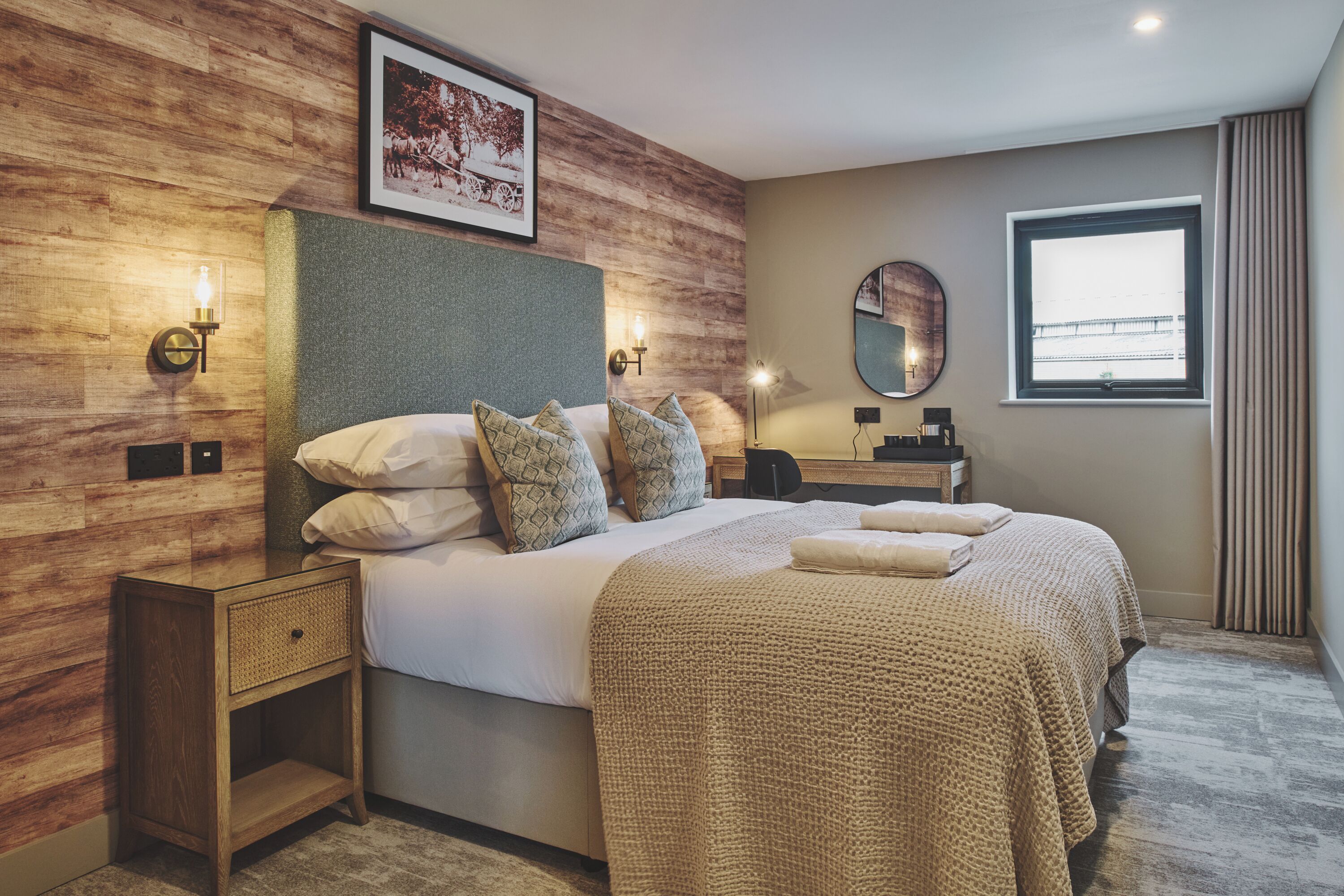 Ein modernes Hotelzimmer mit einem großen Bett, Wandverkleidung aus Holz, einem runden Spiegel und einem Fenster mit Vorhängen.
