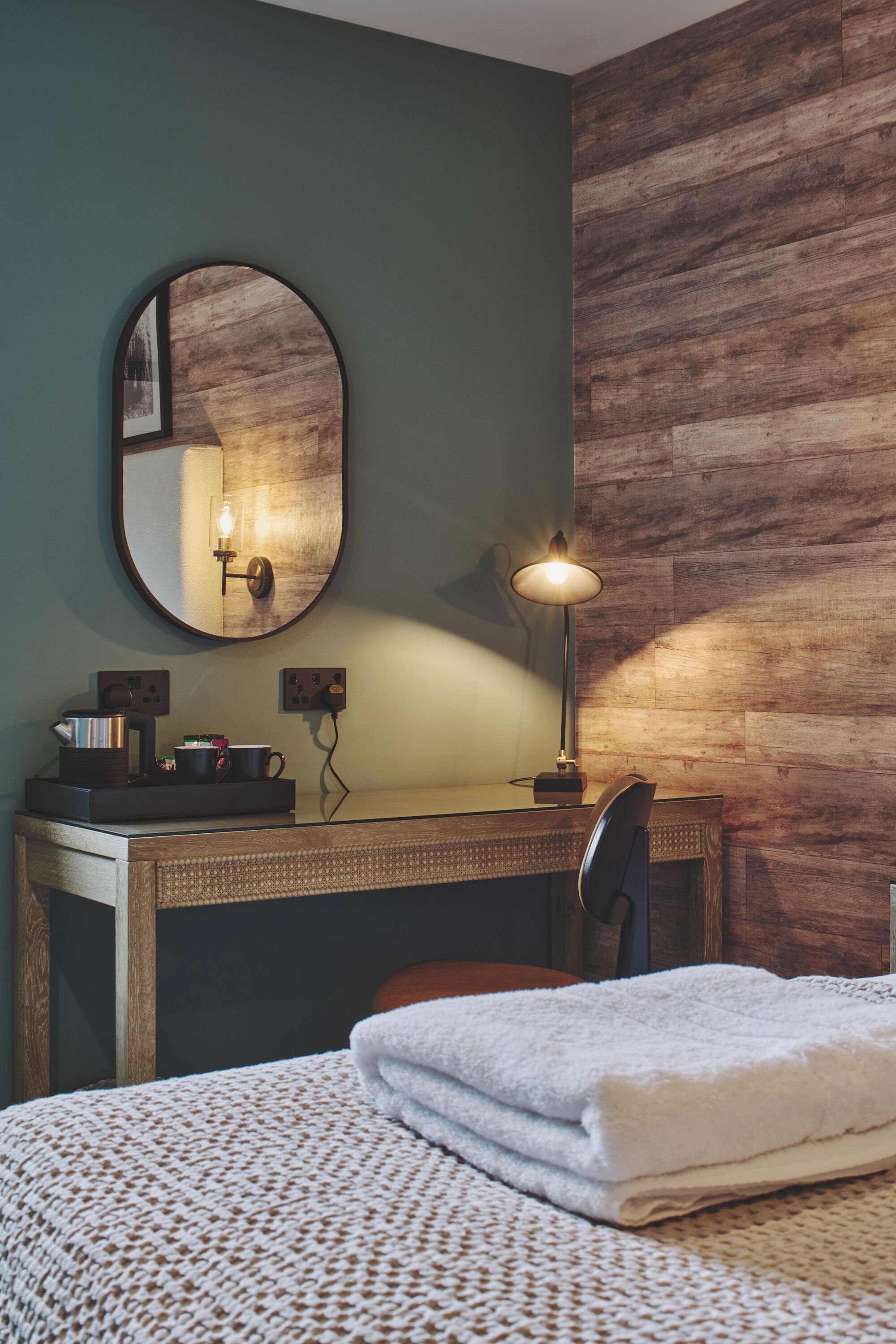 A hotel room with a wooden desk, a round mirror, a lamp, a chair, and a neatly made bed with a white towel.