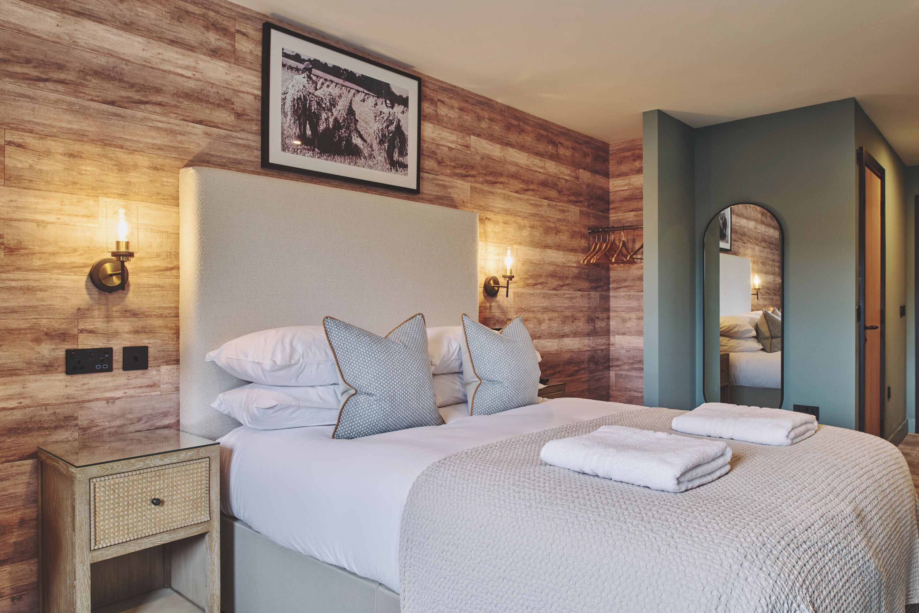 Ein ordentlich eingerichtetes Schlafzimmer mit einem großen Bett, zwei Wandlampen, einem gerahmten Bild über dem Bett und einem Spiegel an der Wand.