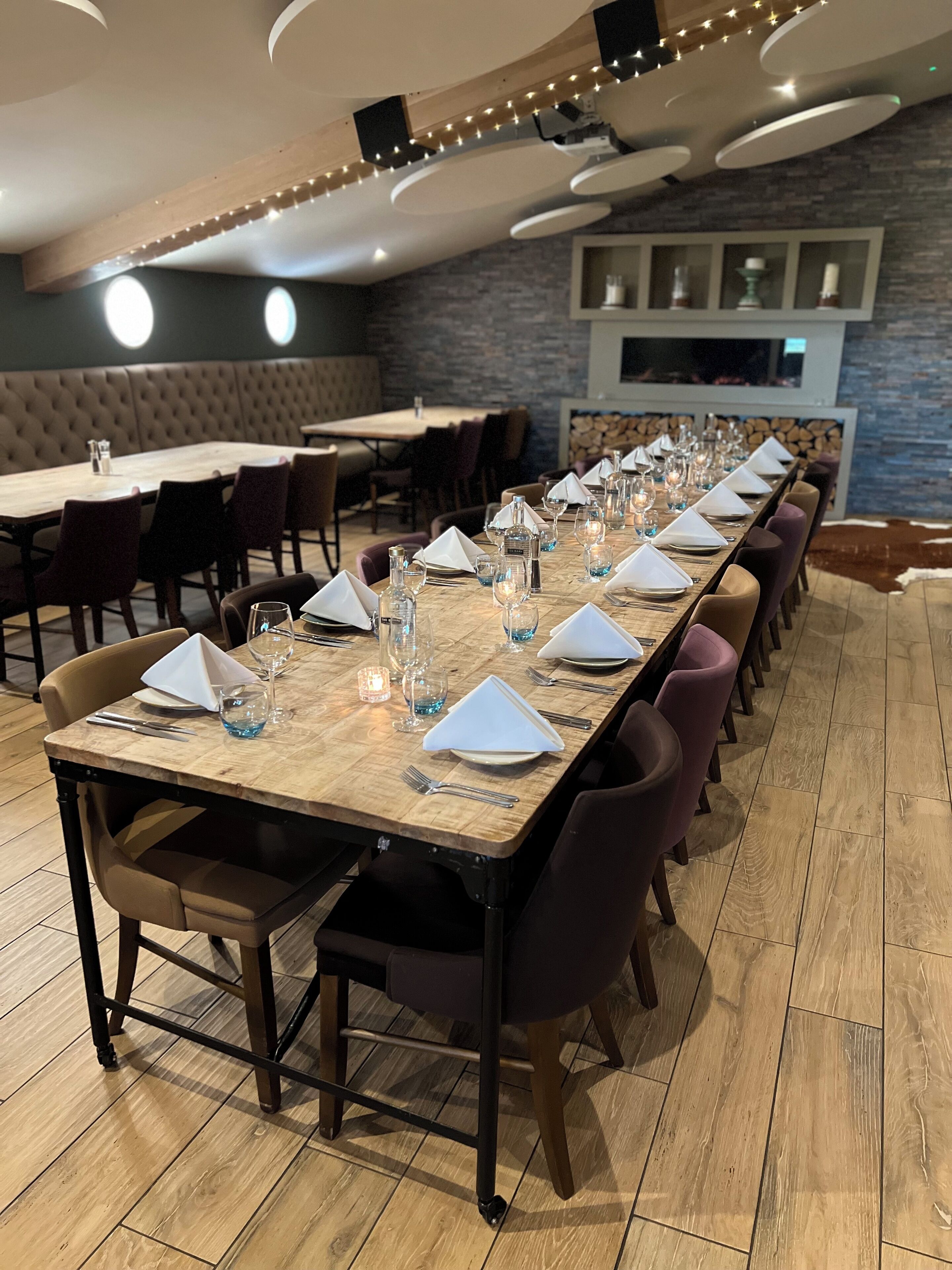 A long dining table set with chairs, candles, and glassware in a restaurant with a brick wall and wooden ceiling.