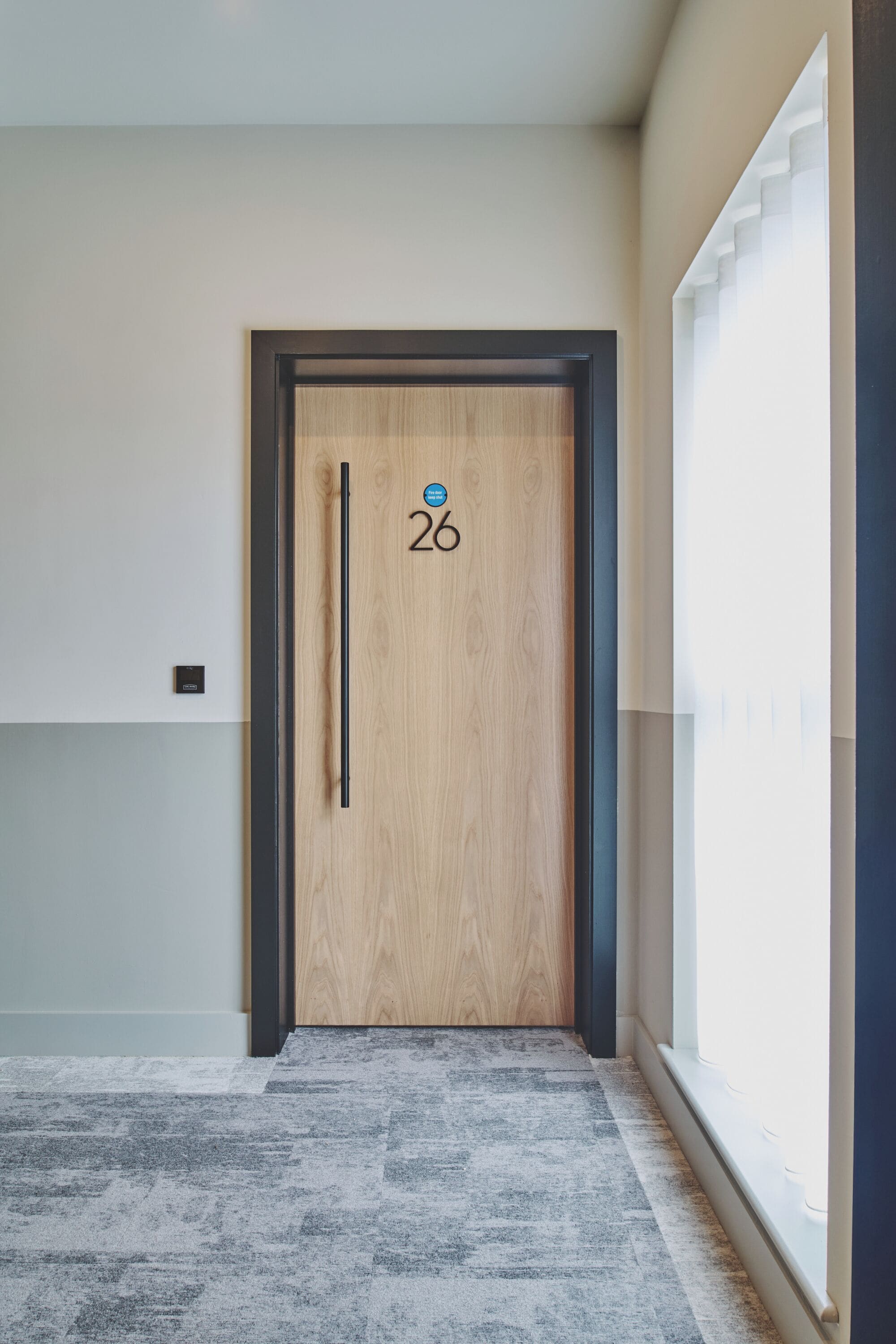 Een gang met een houten deur met het nummer 26, een raam met zonwering en een grijs vloerkleed.