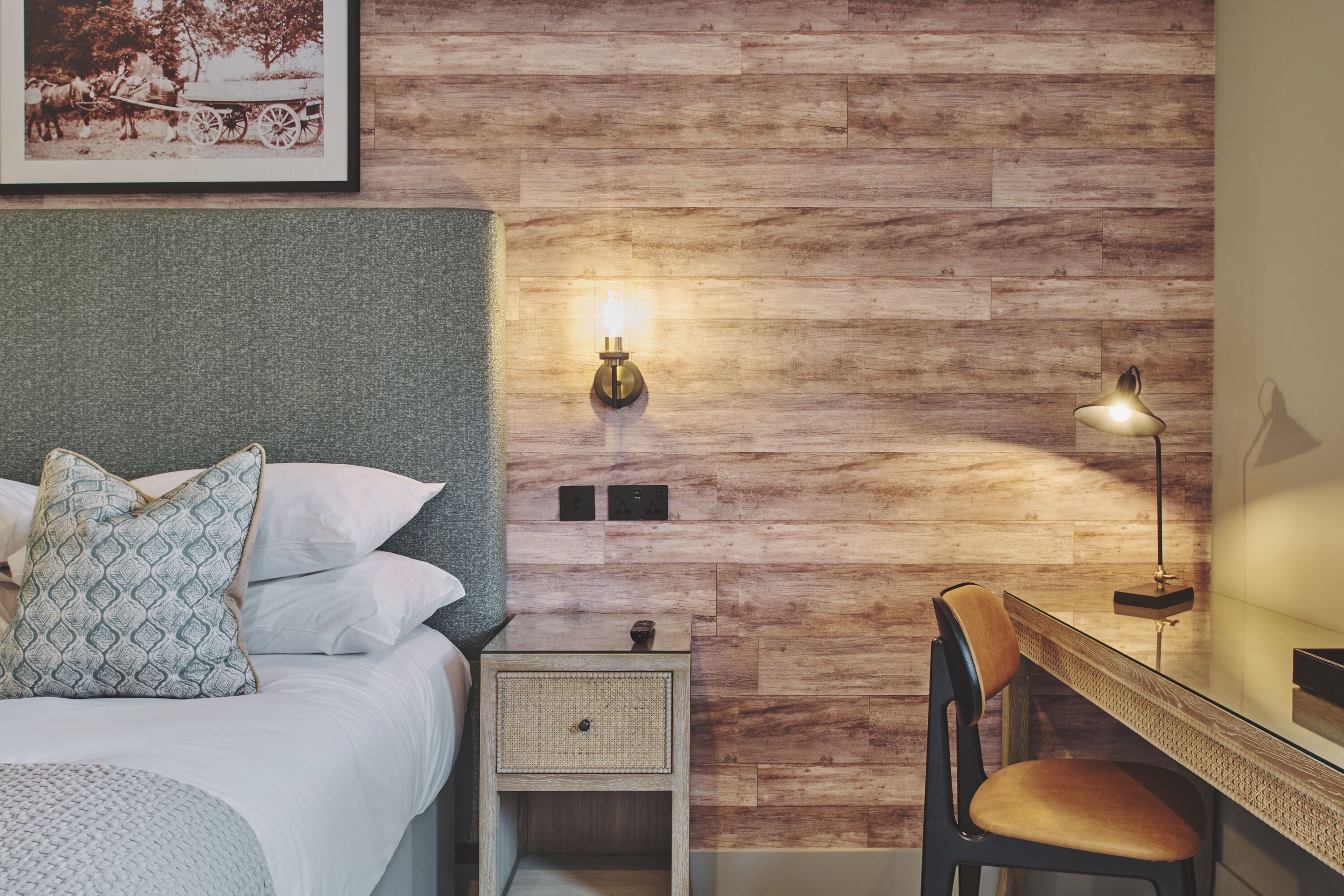 Een slaapkamer met een houten wand, een bed met witte lakens, een nachtkastje met een lamp en een bureau met een stoel.
