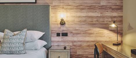 Een slaapkamer met een houten wand, een bed met witte lakens, een nachtkastje met een lamp en een bureau met een stoel.