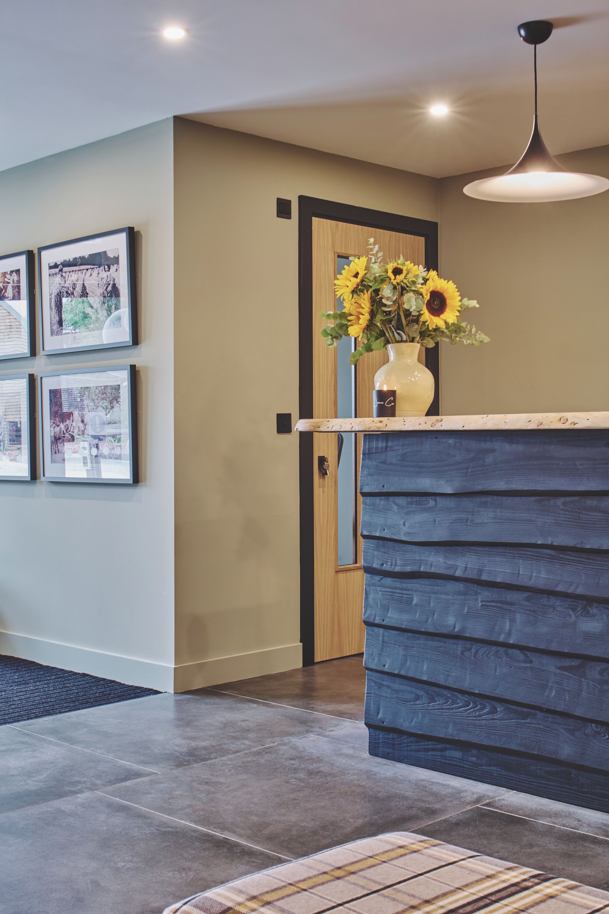 A reception area with a wooden door, a vase of sunflowers, framed pictures on the wall, and a modern pendant light.
