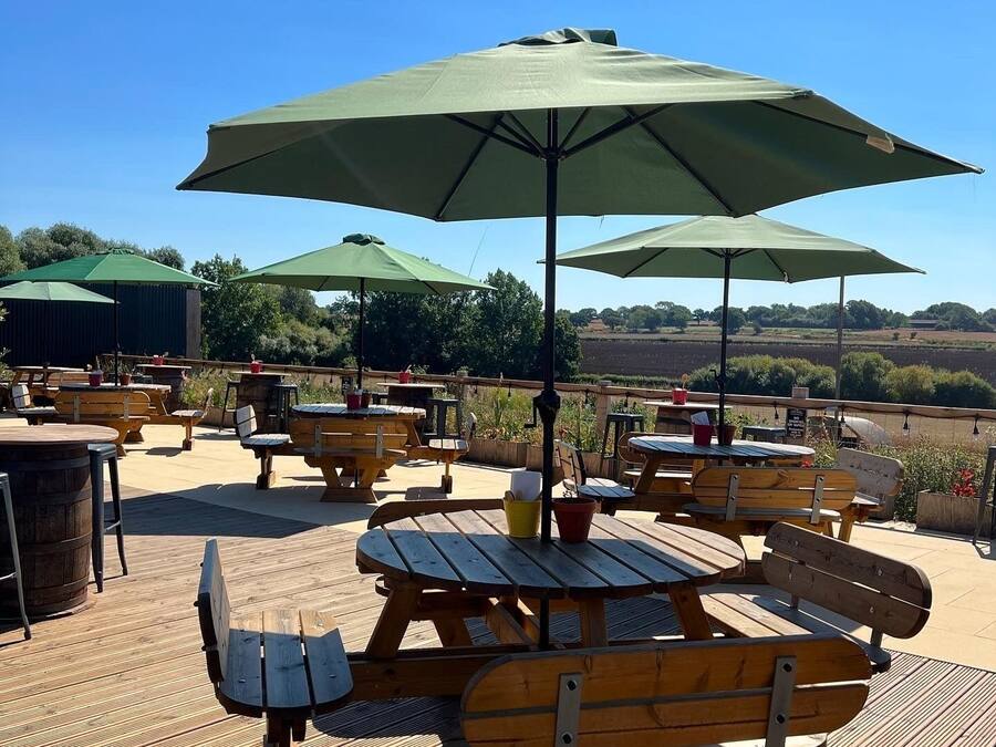 Un patio al aire libre con mesas y bancos de madera, sombrillas verdes y vistas al campo.
