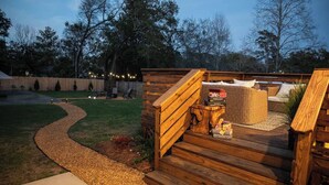 Una terraza de madera con un sendero curvo que lleva a una zona de estar con muebles de mimbre y una mesa pequeña con aperitivos.