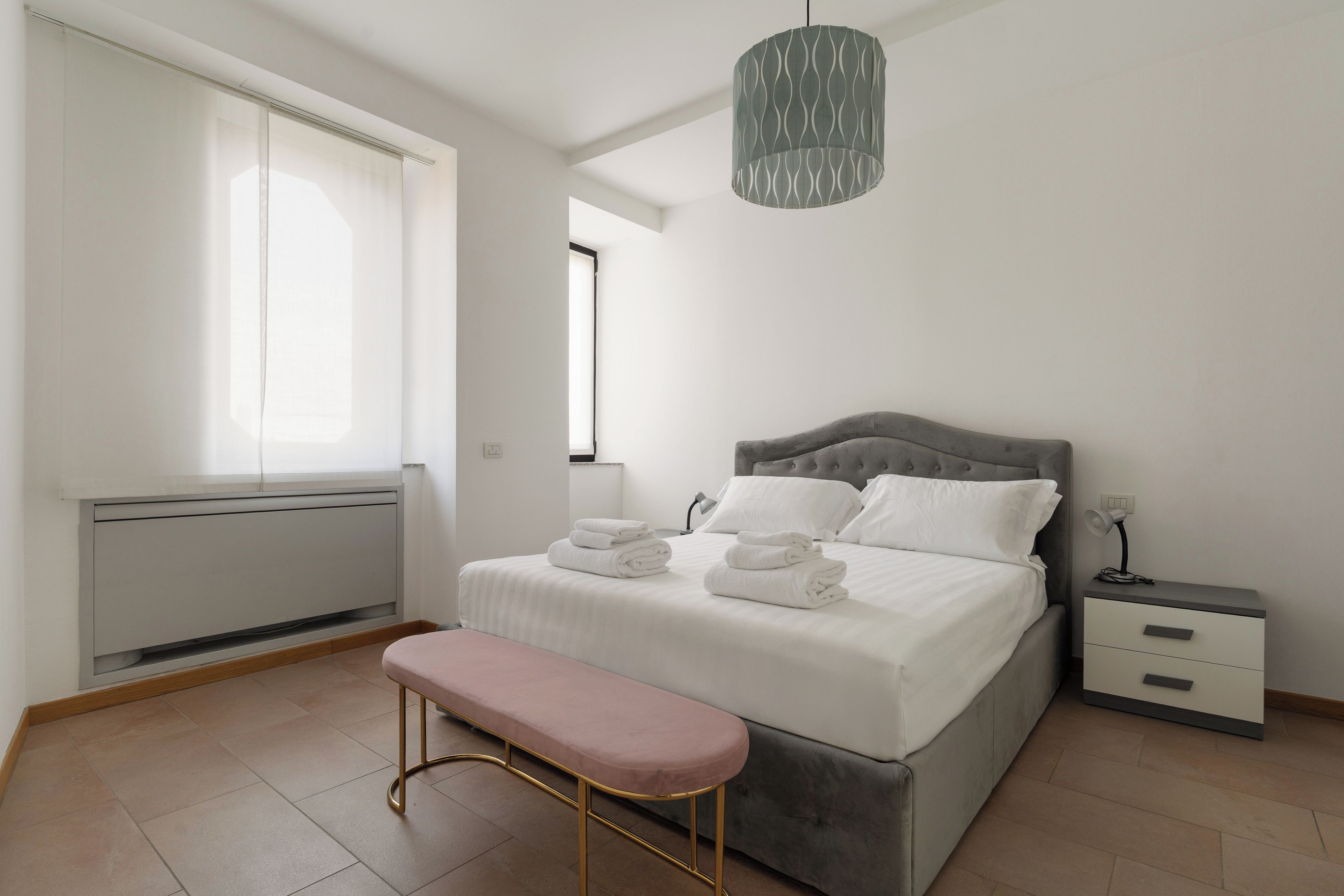 A bedroom with a bed, bedside tables, a bench, and a hanging light fixture.