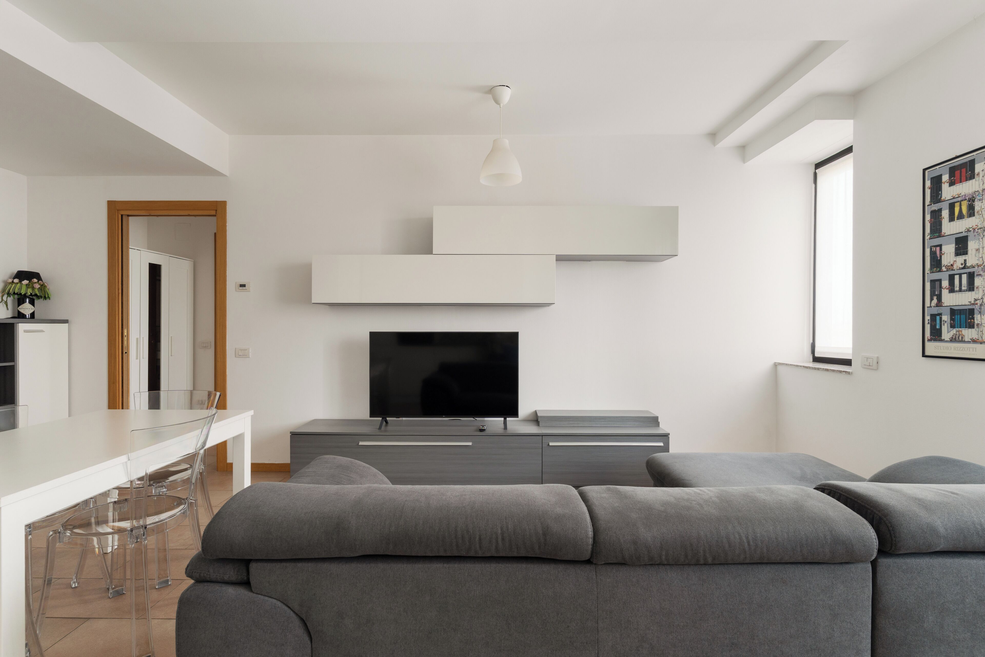 Ein modernes Wohnzimmer mit einem Flachbildfernseher, grauen Sofas, einem Essbereich mit weißem Tisch und einer Küche im Hintergrund.