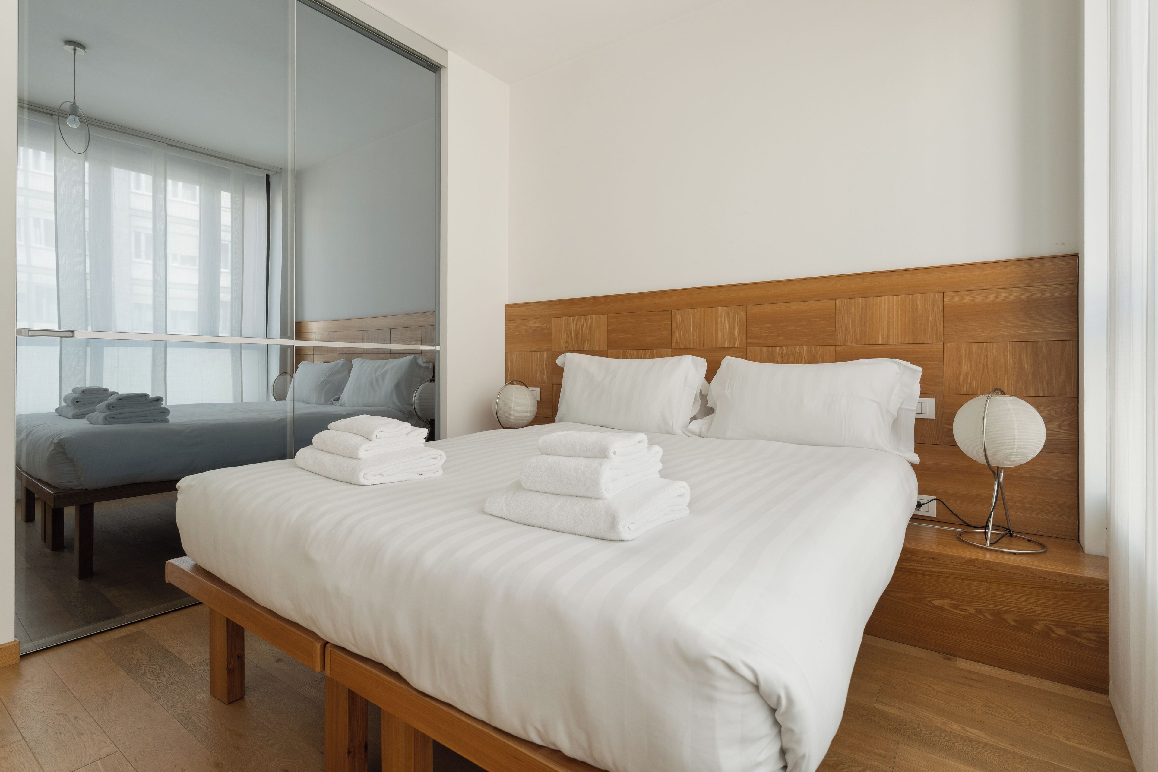 A hotel room with a large bed, a bedside table with a lamp, and a view of the city through a window.