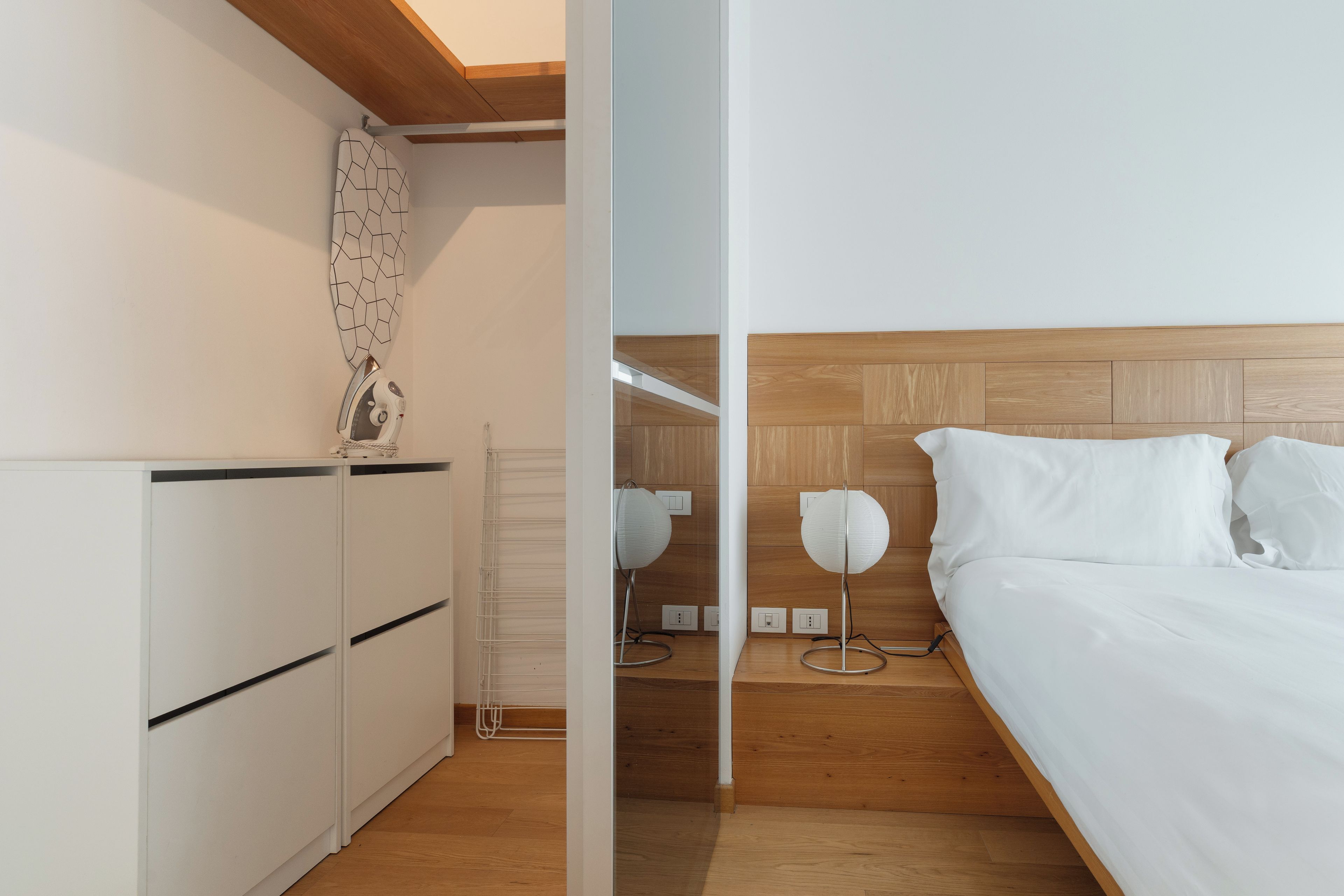 A modern bedroom with a wooden headboard, a bed with white linens, a bedside table with a lamp, and a wardrobe.