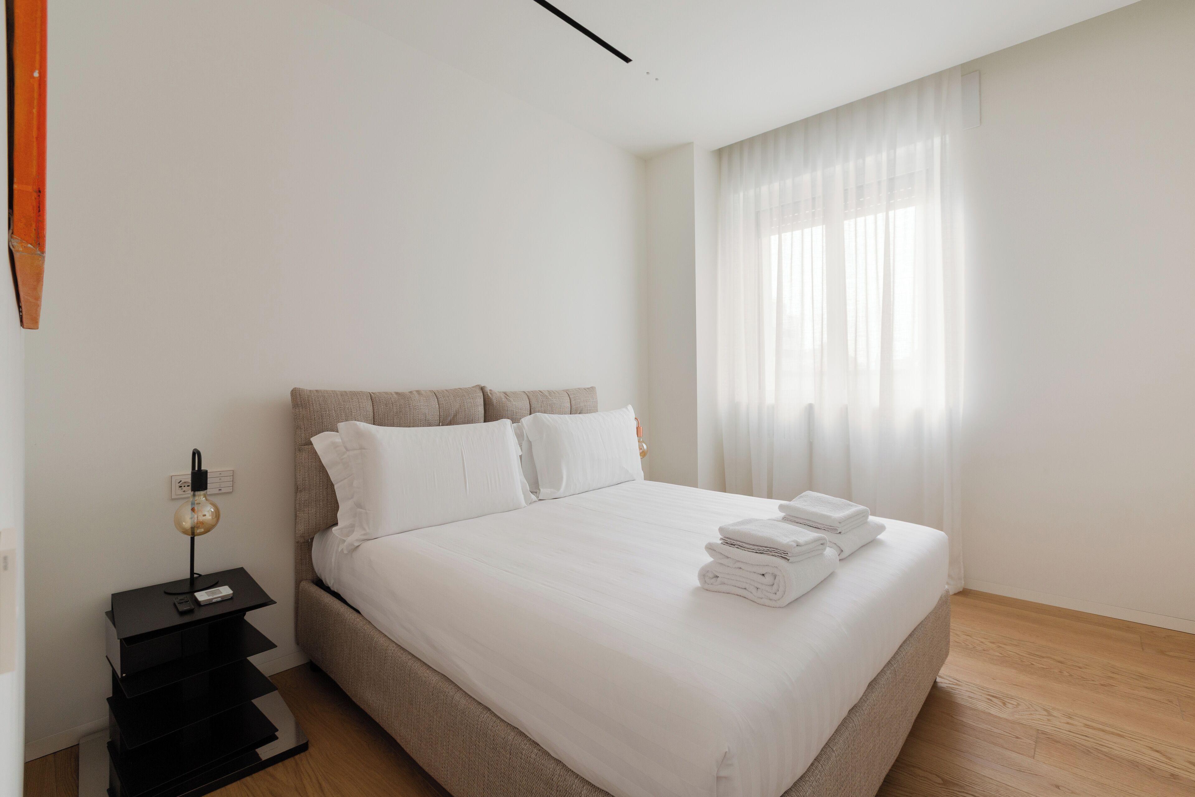 Een netjes opgemaakt bed met witte lakens en een gevouwen handdoek aan de zijkant, een zwart nachtkastje met een lamp, en een raam met dunne gordijnen waardoor natuurlijk licht binnenkomt.