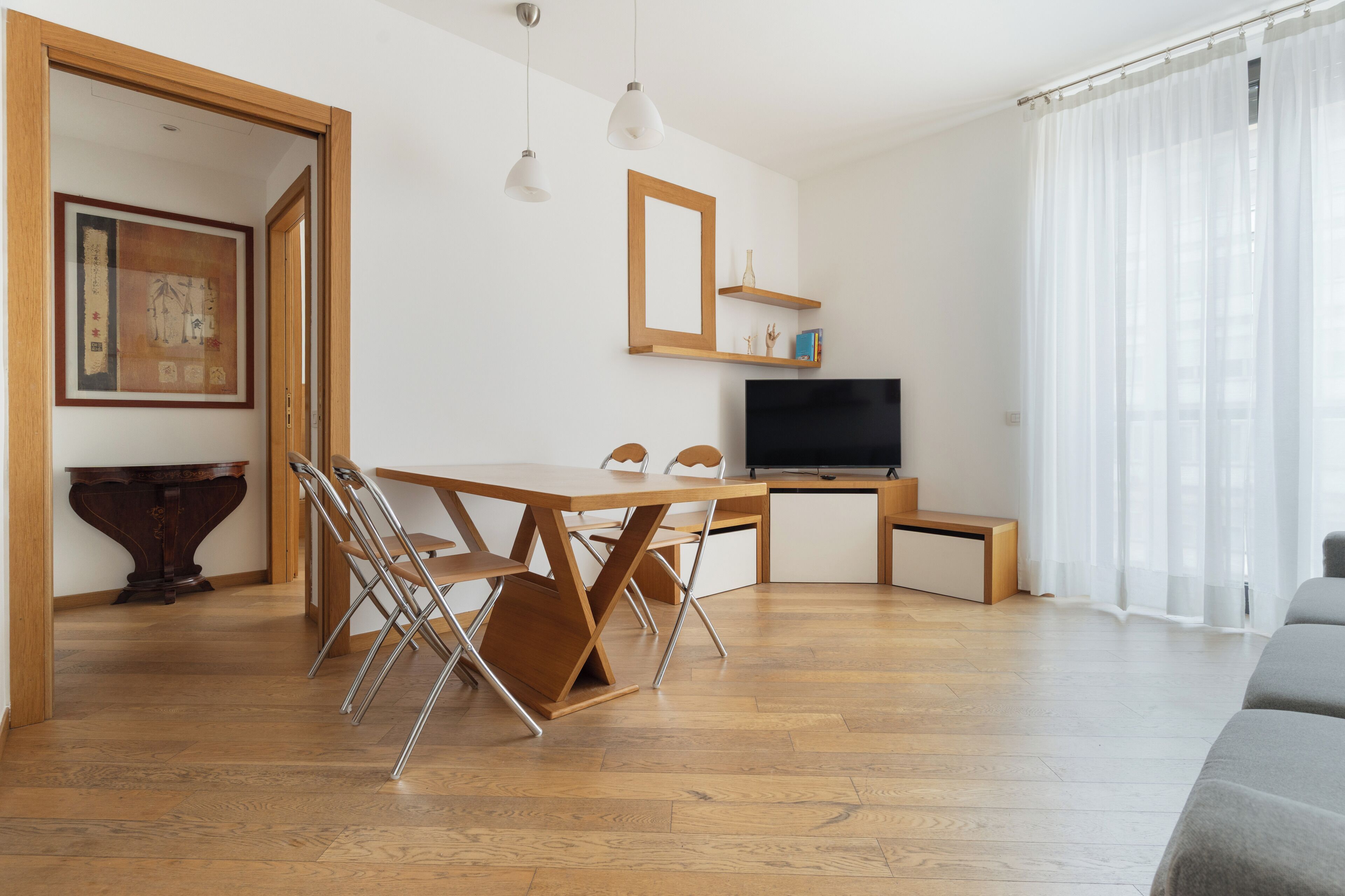 Una sala de estar moderna con una mesa de comedor de madera, sillas, un televisor de pantalla plana y un ventanal con cortinas translúcidas.