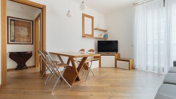 Una sala de estar moderna con una mesa de comedor de madera, sillas, un televisor de pantalla plana y un ventanal con cortinas translúcidas.