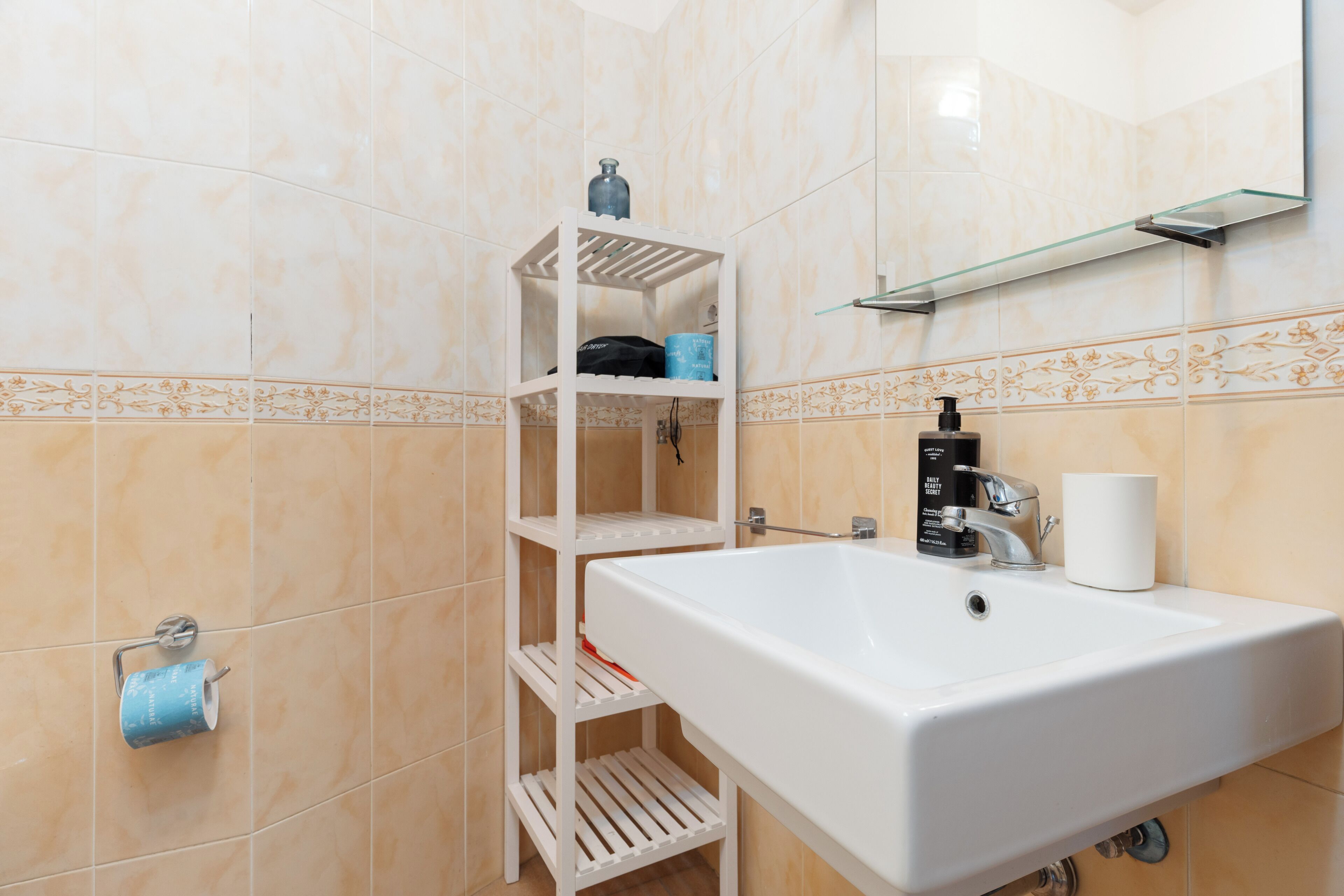 Un baño con un lavamanos blanco, un espejo, un perchero para toallas y un estante con artículos de aseo.