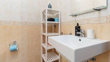 Un baño con un lavamanos blanco, un espejo, un perchero para toallas y un estante con artículos de aseo.
