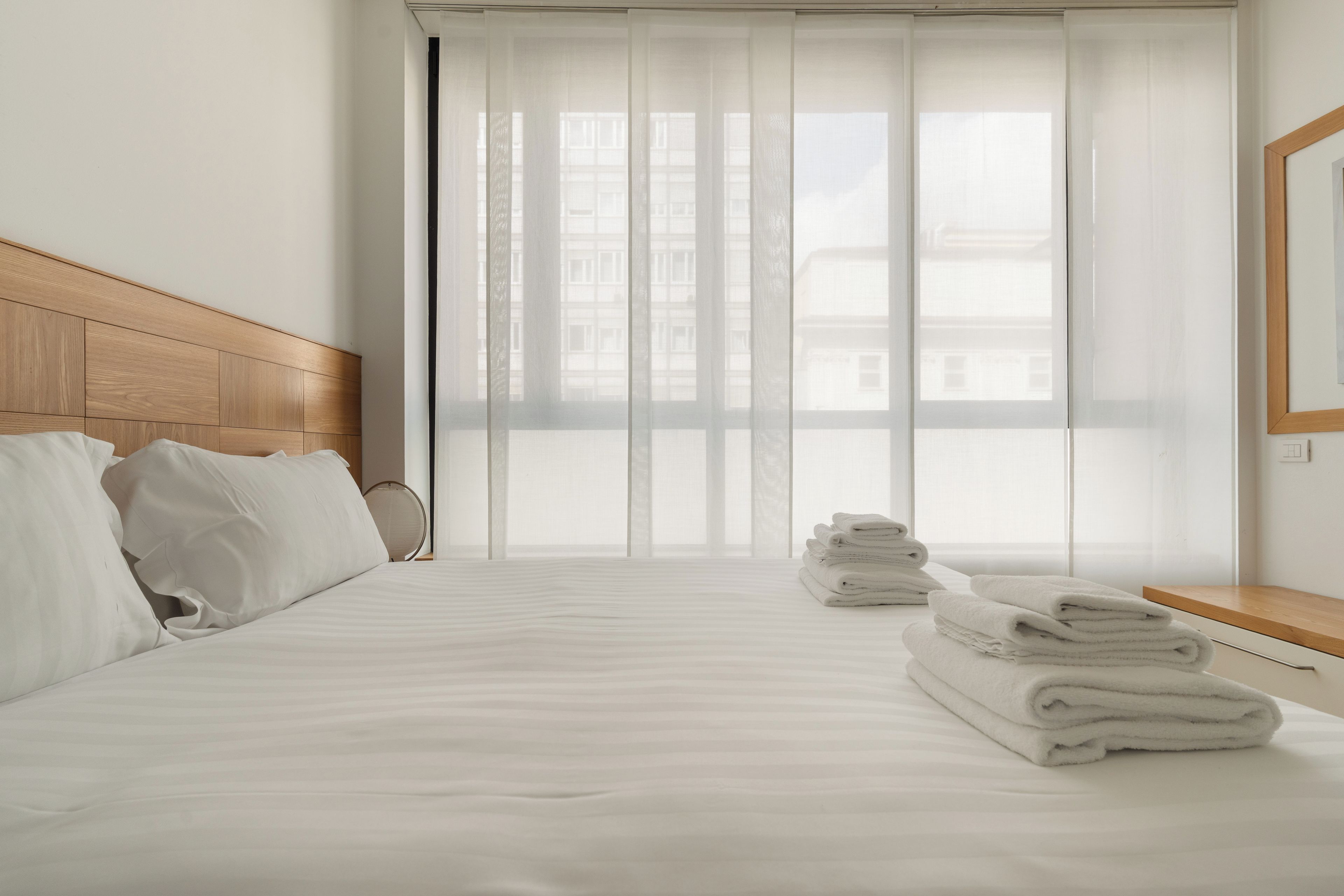 Habitación de hotel con cama, cabecera de madera, ventana con cortinas traslúcidas y una pila de toallas sobre una mesita de noche de madera.