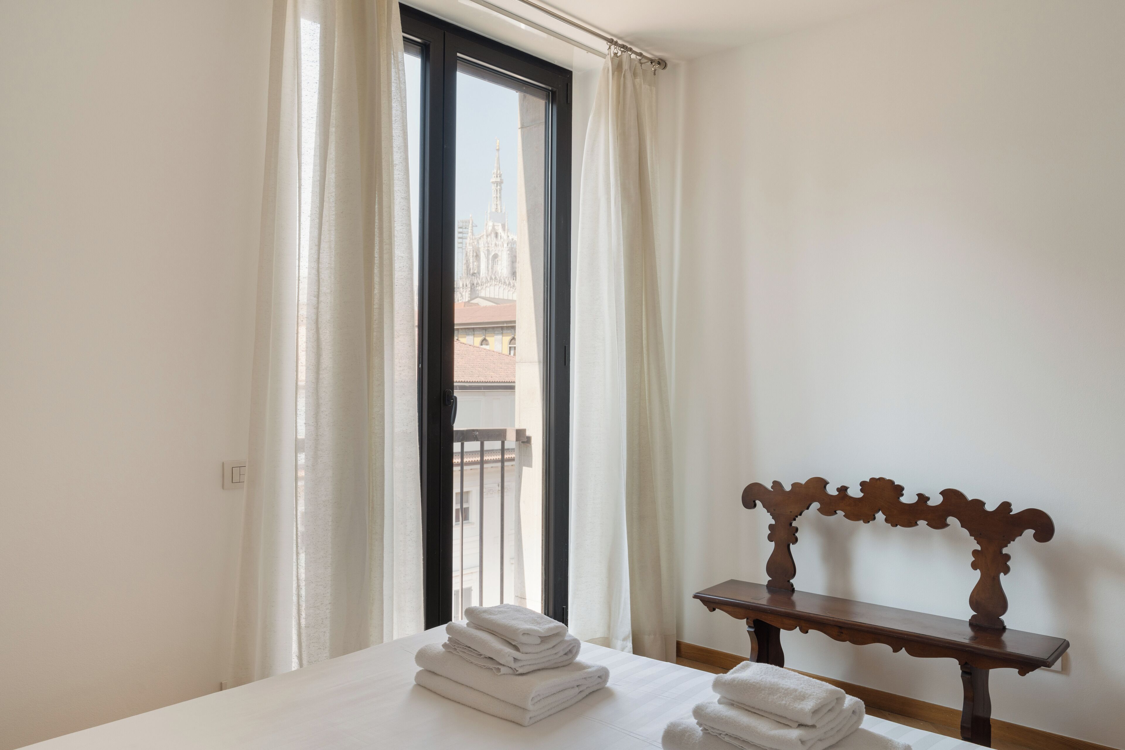 Une chambre d’hôtel avec un lit, un banc en bois et une vue sur un immeuble par la fenêtre.