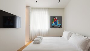 Ein modernes Hotelzimmer mit einem großen Bett, einem an der Wand befestigten Flachbildfernseher und einem Gemälde, das ein Paar auf einem Roller zeigt.