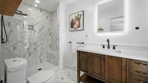 Une salle de bain moderne avec un mur de douche en marbre, un lavabo blanc doté d’un meuble-vasque en bois et une estampe botanique encadrée au mur.