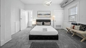 Une chambre avec un grand lit, une table de chevet, un banc avec des coussins, un tableau au mur et un luminaire au plafond.