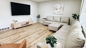 A modern living room with a wooden floor, a beige sofa, a small table with a plant, and a flat-screen TV mounted on the wall. - Newly Renovated with heated Pool, close to CWS,
sleeps 12 (Omaha)