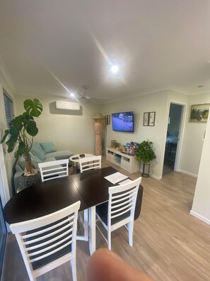 Een zithoek met een ronde tafel, witte stoelen, een potplant, een televisie en een bank.