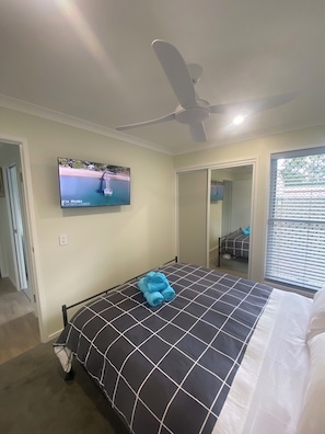 Een slaapkamer met een bed, een plafondventilator, een televisie aan de muur en een raam met jaloezieën.