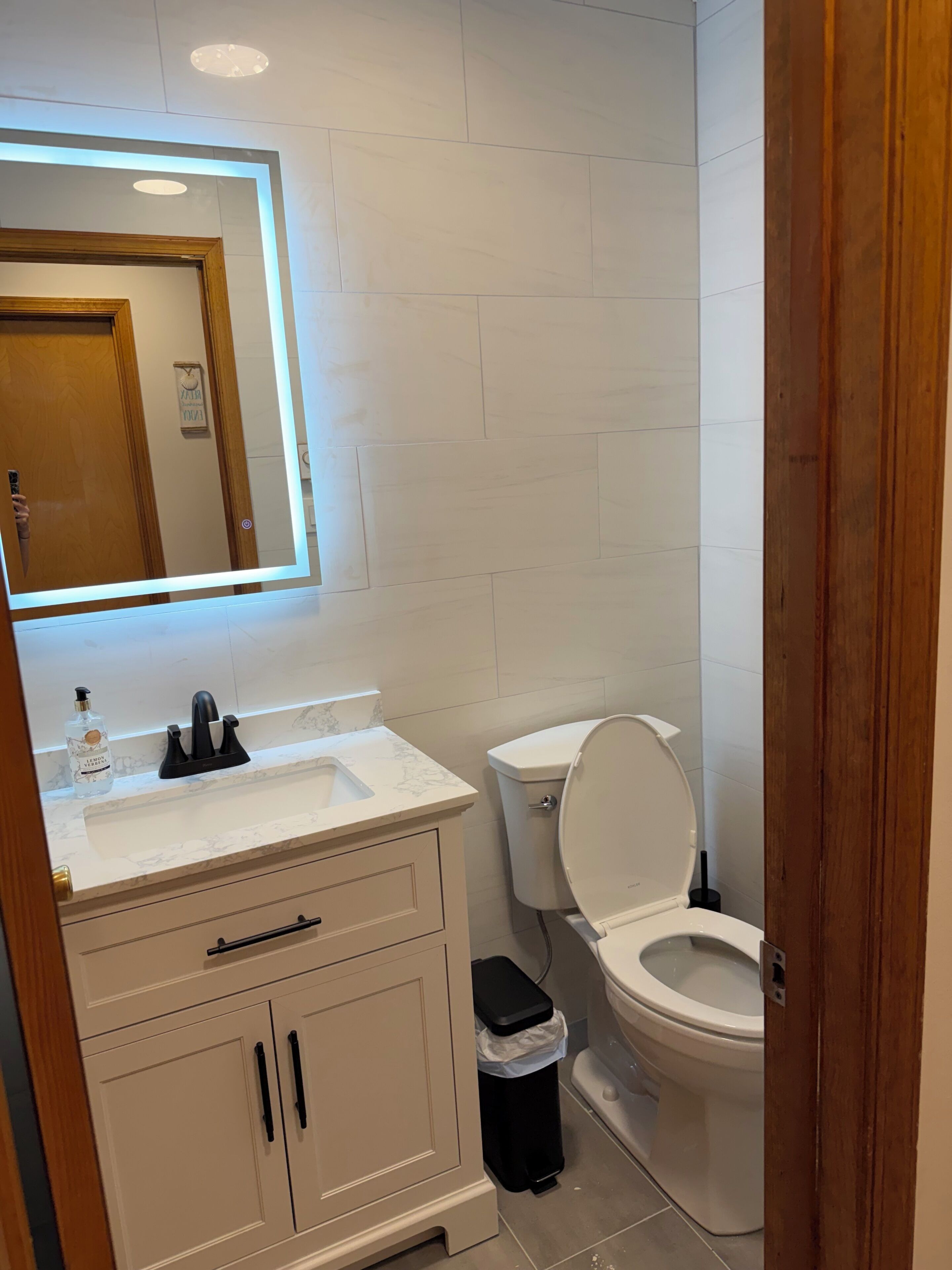 Een badkamer met een wit wastafelmeubel, een spiegel met verlichting, een toilet en een prullenbak.
