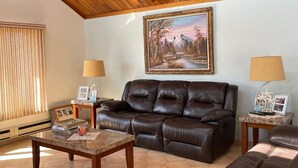 Ein gemütliches Wohnzimmer mit Holzbalkendecke, einem braunen Ledersofa, zwei Beistelltischen mit Lampen und einem gerahmten Gemälde an der Wand.