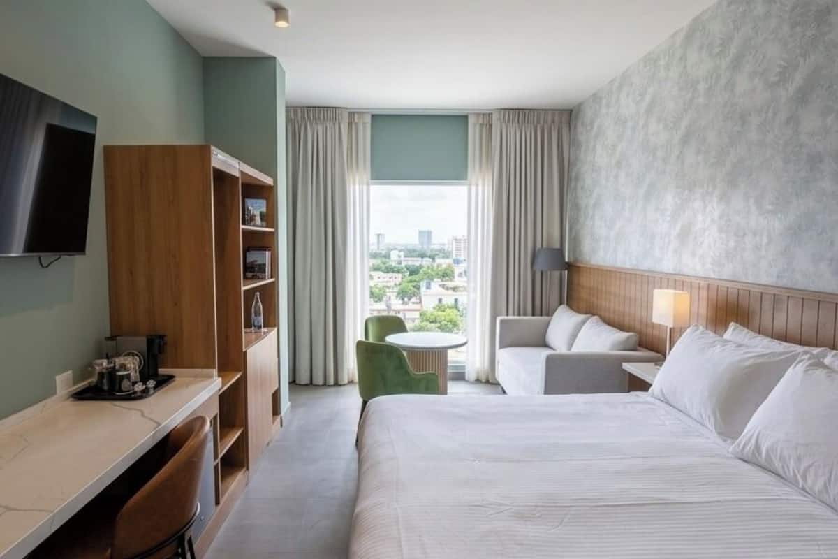 A hotel room with a bed, a desk, a chair, a TV, a bookshelf, and a view of the city.