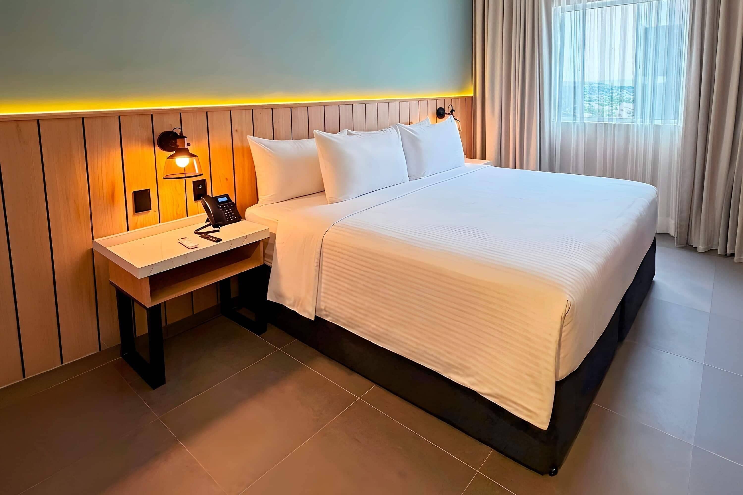 A hotel room with a bed, a bedside table with a lamp, a phone, and a window with curtains.