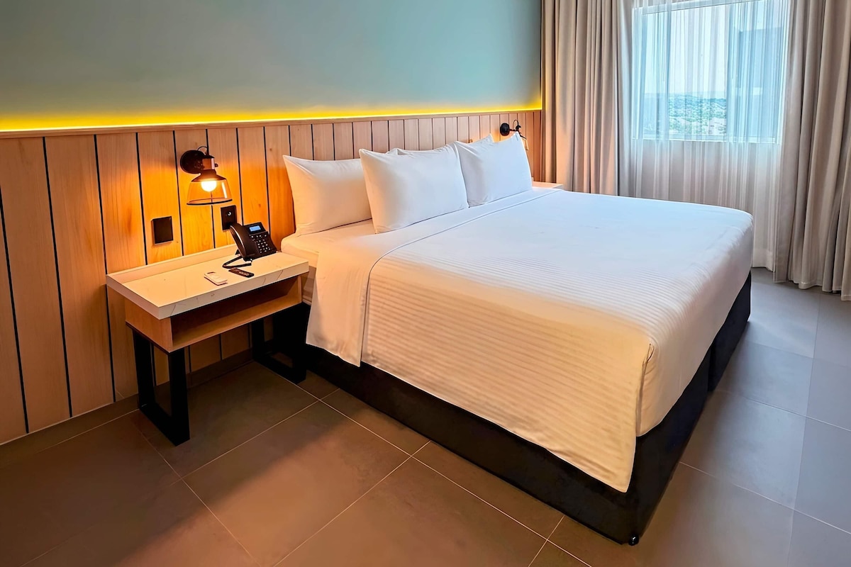 A hotel room with a bed, a bedside table with a lamp, a phone, and a window with curtains.