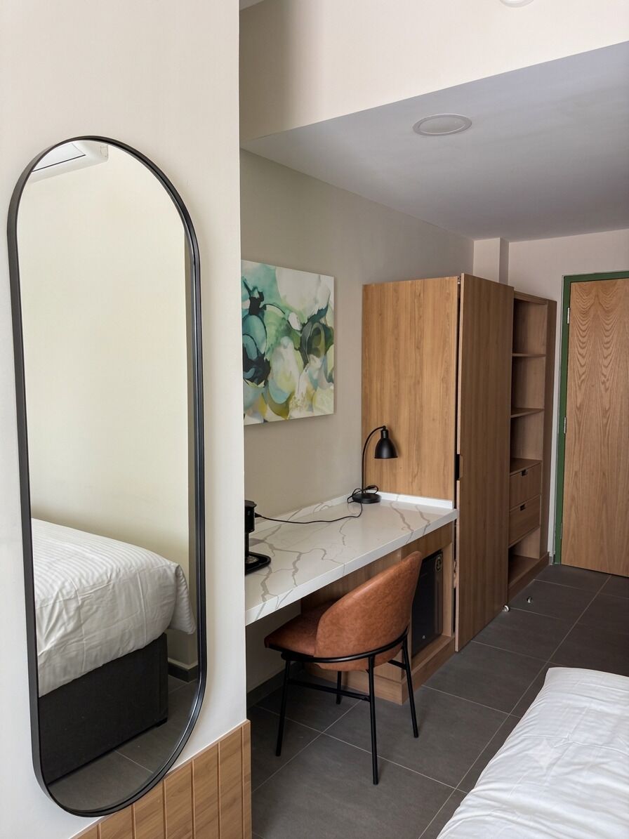 A bedroom with a bed, a desk with a chair, a mirror, a painting, and a wardrobe.