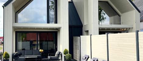 A modern house with a sloped roof, a patio with a sofa and chairs, and a row of sun loungers.
