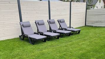A row of outdoor lounge chairs on a well-maintained lawn with a low wall and a tree in the background.