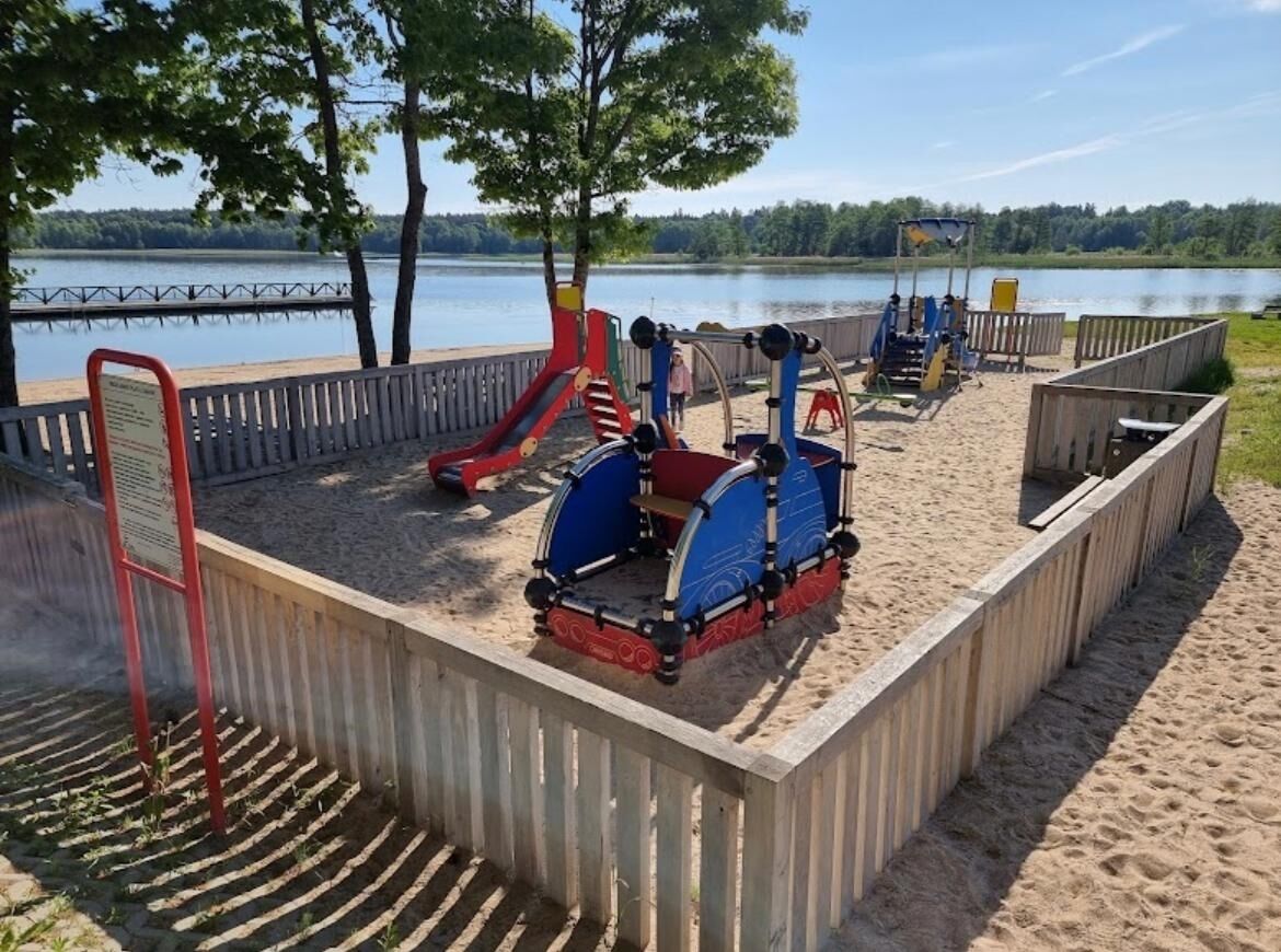 Un parque infantil con diversas estructuras de juego, incluyendo toboganes y juegos de escalar, rodeado por una valla de madera y con vistas a un lago y árboles.