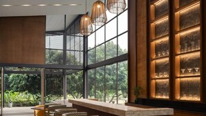 A modern bar area with a wooden counter, cushioned seating, and large windows offering a view of greenery.