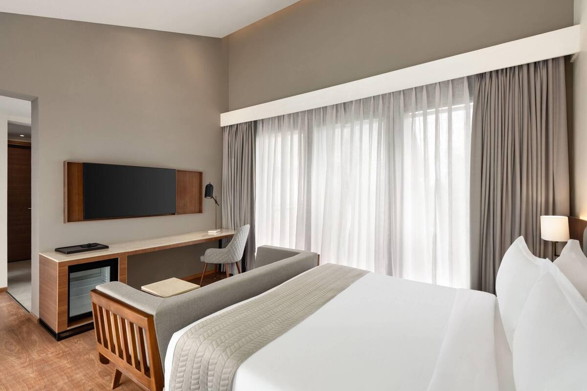 A hotel room with a large bed, a desk with a chair, a TV mounted on the wall, and a window with curtains.