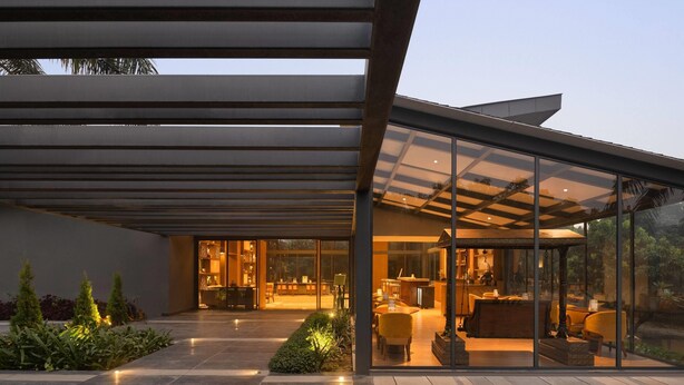 A modern house with a glass facade, outdoor seating area, and a well-lit interior.