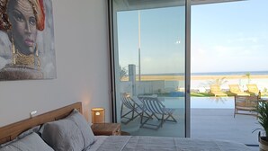 A bedroom with a large bed, a painting on the wall, and a view of a pool and beach through a sliding door.