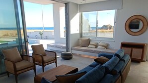 A modern living room with a large window overlooking a beach, a sofa, chairs, and a small table.