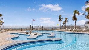 A swimming pool with a curved design, surrounded by lounge chairs and palm trees, with an American flag visible. - Crystal Tower 1703 (Gulf Shores)