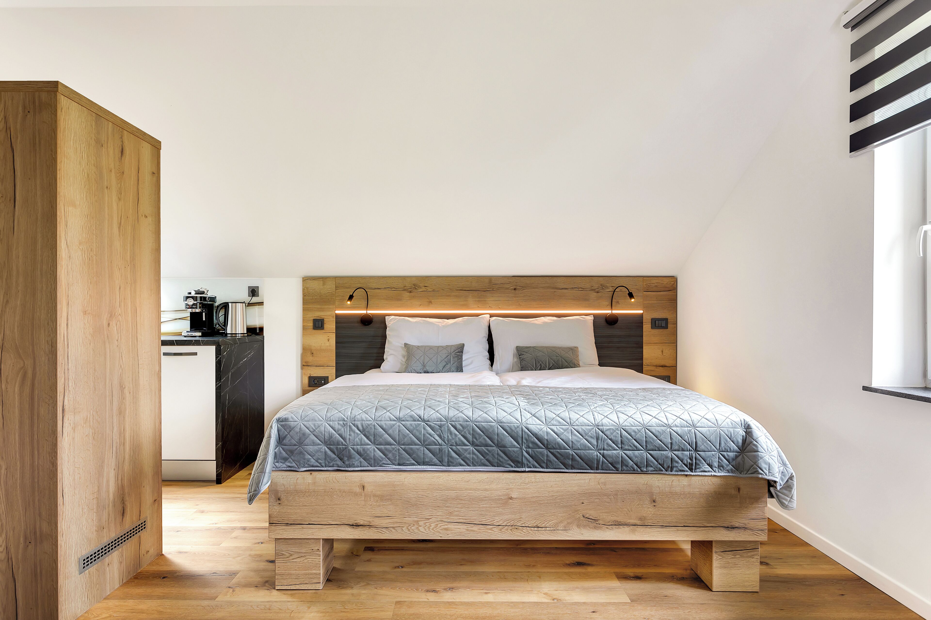 A bedroom with a wooden headboard, a bed with a quilted grey bedspread, and a small kitchenette in the background.