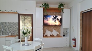 Ein Essbereich mit einem runden Tisch, Stühlen, einer Blumenvase, einem an der Wand befestigten Fernseher und einem Feuerlöscher.