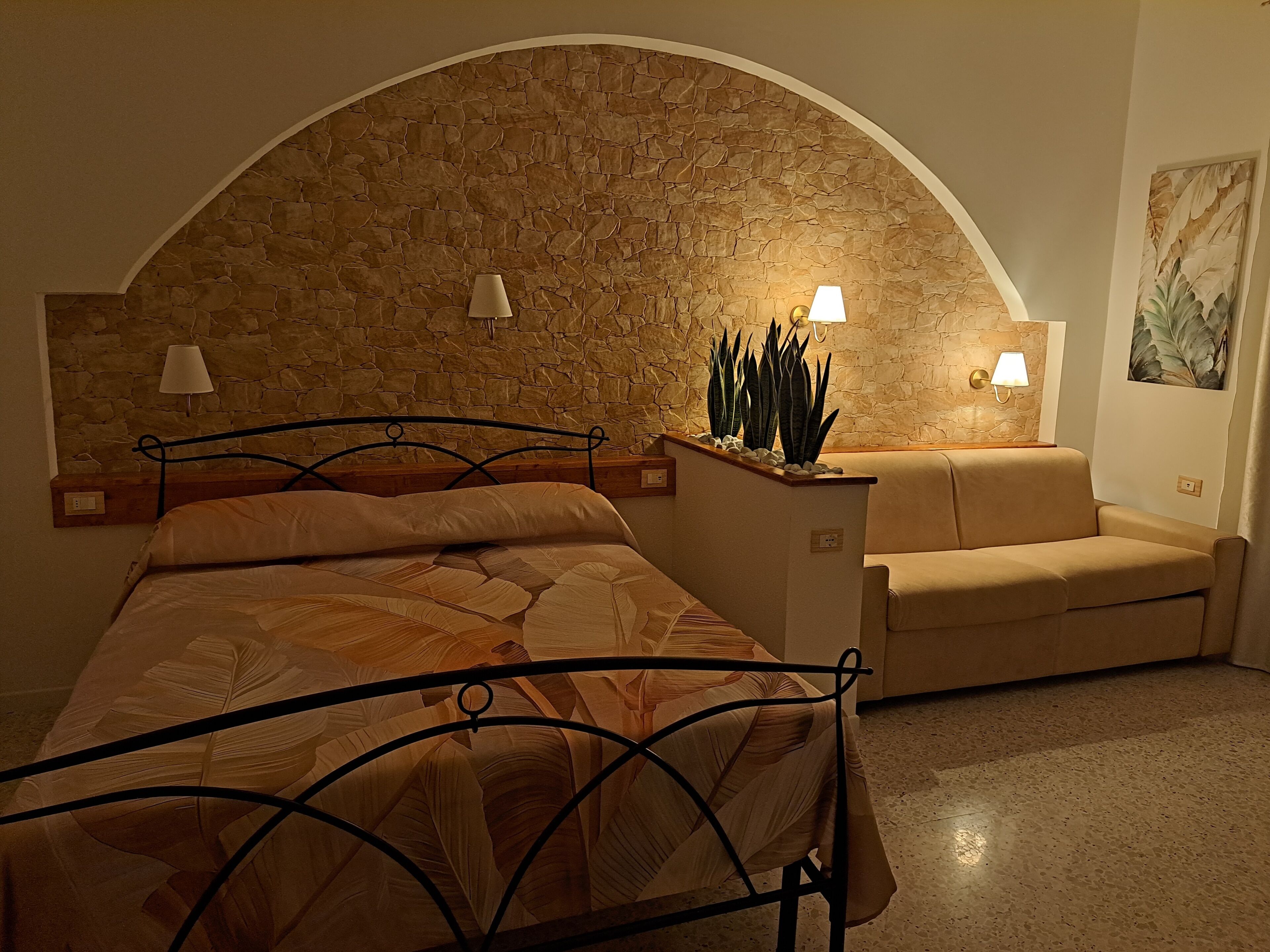 A bedroom with a stone wall, a bed with a leaf-patterned bedspread, a nightstand, a lamp, and a small table with plants.